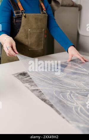 Zugeschnittene Aufnahme einer jungen Handwerkerin in Schürze, die im Typografie-Zentrum neben der Werkbank steht und handgefertigte Kunstwerke auf Skizze setzt Stockfoto