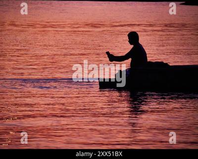 Silhouette eines Mannes, der bei Sonnenuntergang am Wasser sitzt Stockfoto