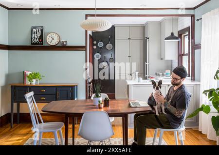 Hund sitzt auf dem Schoß eines Mannes in einem modernisierten Esszimmer mit Blick in die renovierte kleine Küche Stockfoto