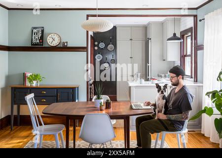 Hund sitzt auf dem Schoß eines Mannes in einem modernisierten Esszimmer mit Blick in die renovierte kleine Küche Stockfoto