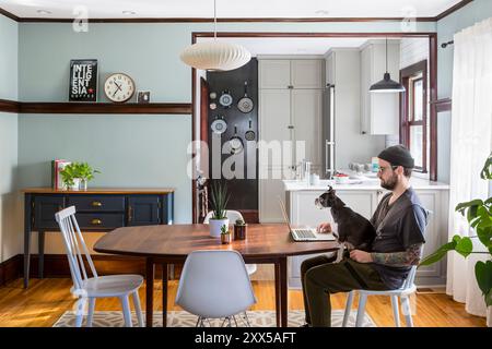 Hund sitzt auf dem Schoß eines Mannes in einem modernisierten Esszimmer mit Blick in die renovierte kleine Küche Stockfoto