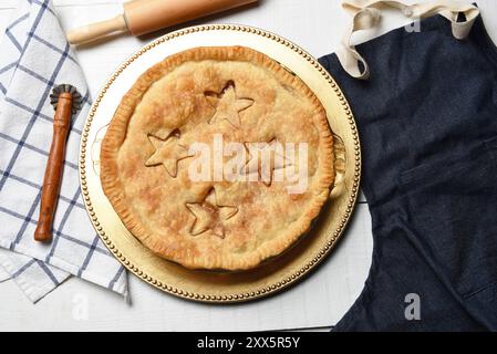 Frisch gebackener hausgemachter Apfelkuchen auf einem weißen Holztisch mit Schürze, Nudelnadel und Crimper. Stockfoto