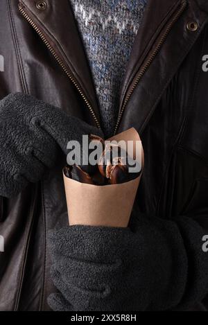 Nahaufnahme eines Mannes in Pullover und Lederjacke, der einen braunen Papierkegel mit frisch gerösteten Kastanien hält. Stockfoto