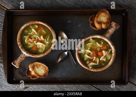 Zwei Schüsseln Chicken Nudelsuppe, ein Baguette Knoblauch Toast und Löffel auf einem Blech auf einem rustikalen Holzküchentisch. Stockfoto