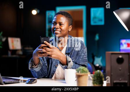 Während sie an ihrem Schreibtisch sitzt, überprüft afroamerikanische Selbstständige das Handy nach Nachrichten. Eine junge schwarze Frau, die ein Mobilgerät benutzt, um in sozialen Medien zu chatten. Stockfoto