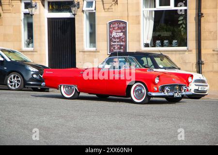 Ein klassisches rotes Cabriolet ford thunderbird, das eine Straße in der Nähe eines Cafés entlang fährt, mit anderen geparkten Autos und einem Gebäude im Hintergrund. Stockfoto