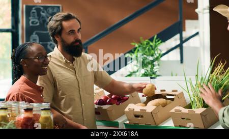 Händler zeigt reifes Gemüse an Kunden, die Lebensmittel einkaufen gehen, verschiedene Paare wählen Kartoffeln und veganes Essen für gesunde Mahlzeiten. Lokaler Anbieter, der Bioprodukte empfiehlt. Handgeführte Aufnahme. Stockfoto