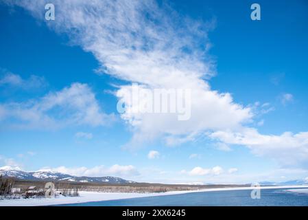 Ein blauer, heller Tag mit teilweise bewölkten Wolken und einem großen Fluss mit schneebedeckten Bergen und schneebedeckter Landschaft in Tagish, Yukon, Kanada Stockfoto