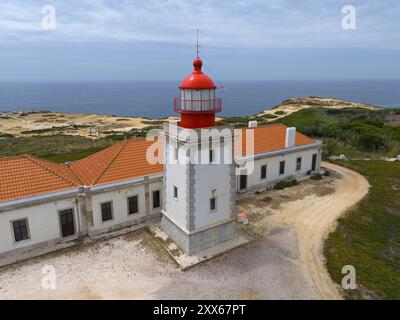 Nahaufnahme eines Leuchtturms und eines benachbarten Gebäudes mit Blick auf das Meer unter bewölktem Himmel aus der Luft, Farol do Cabo Sardao, Sardao, Ponta do Caval Stockfoto