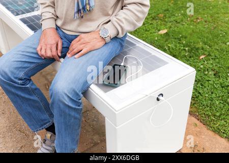 Der Mann sitzt auf einer Smart Bench und lädt sein Mobiltelefon, moderne Technologie, Bank mit Solarmodul und Handy-Ladegerät auf Stockfoto