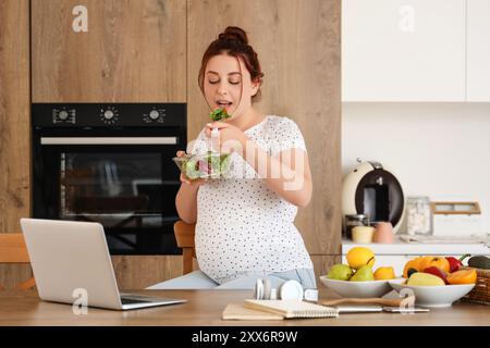 Junge schwangere Frau mit Laptop, die frischen Gemüsesalat in der Küche isst Stockfoto