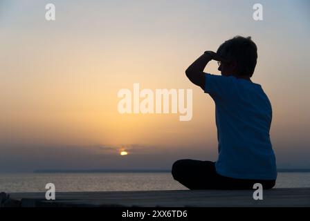 Frau sitzt am Ufer des Mittelmeers und blickt in die untergehende Sonne Stockfoto