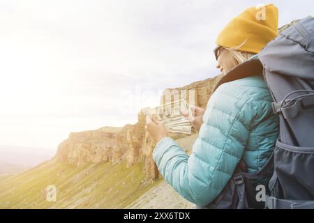 Ein Reisender mit Hut und Sonnenbrille hält hundert Dollar-Scheine in den Händen eines Ventilators vor dem Hintergrund von Felsen auf der Natur. Reisekosten Stockfoto