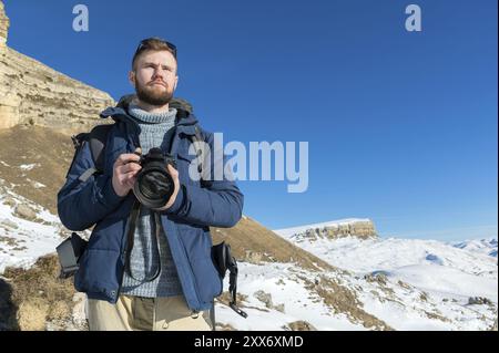 Porträtieren Sie Einen bärtigen Hipster-Fotografen mit Rucksack und Sonnenbrille mit Standfüßen mit einer DSLR-Kamera in den Händen vor dem Hintergrund von e Stockfoto