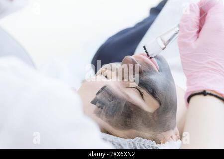 Der Kosmetologe in pinkfarbenen Handschuhen mit Pinsel trägt eine Kohlemaske zum Schälen auf das Gesicht eines jungen Mädchens in einem Kosmetologie-Raum auf. Das Konzept des Kosms Stockfoto