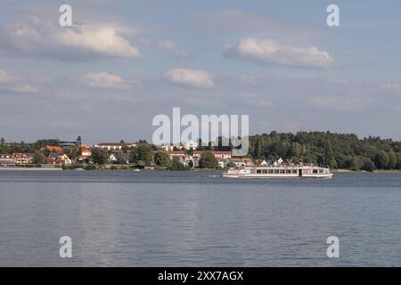 Ein weißes Ausflugsboot gleitet auf dem Müritz-See vor der Stadt Waren, Deutschland, mit farbenfrohen Gebäuden und üppigem Grün, unter hellblauem Himmel Stockfoto