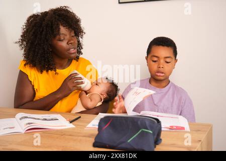 Junge Mutter ernährt Neugeborene und hilft dem Sohn bei Hausaufgaben zu Hause Stockfoto