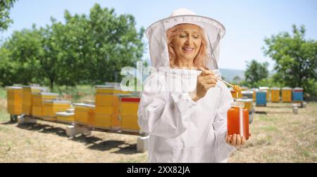 Weibliche Imkerin in Uniform, die Honig aus einem Glas probiert und lächelt Stockfoto