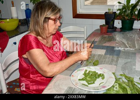 Eine ältere Frau saß an ihrem Küchentisch und Schnitt mit einem Messer vorsichtig grüne Bohnen. Die Szene spiegelt die Einfachheit und den Komfort des täglichen Lebens wider, wh Stockfoto