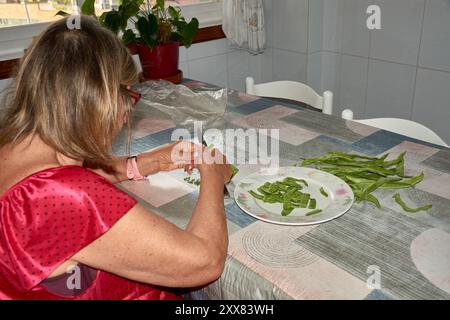 Eine ältere Frau saß an ihrem Küchentisch und Schnitt mit einem Messer vorsichtig grüne Bohnen. Die Szene spiegelt die Einfachheit und den Komfort des täglichen Lebens wider, wh Stockfoto