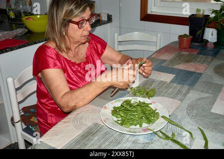 Eine ältere Frau saß an ihrem Küchentisch und Schnitt mit einem Messer vorsichtig grüne Bohnen. Die Szene spiegelt die Einfachheit und den Komfort des täglichen Lebens wider, wh Stockfoto