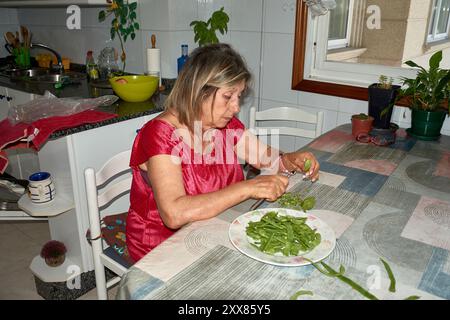 Eine ältere Frau saß an ihrem Küchentisch und Schnitt mit einem Messer vorsichtig grüne Bohnen. Die Szene spiegelt die Einfachheit und den Komfort des täglichen Lebens wider, wh Stockfoto