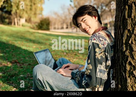 Junger asiatischer Student, der in einem Park sitzt und seinen Laptop benutzt, um zu lernen Stockfoto
