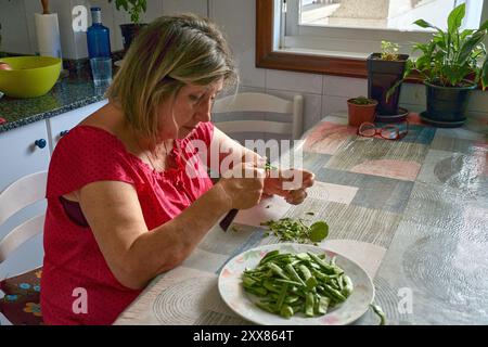 Eine ältere Frau saß an ihrem Küchentisch und Schnitt mit einem Messer vorsichtig grüne Bohnen. Die Szene spiegelt die Einfachheit und den Komfort des täglichen Lebens wider, wh Stockfoto