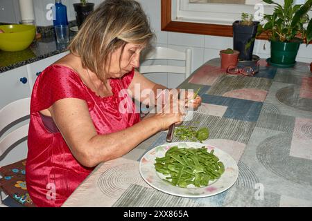 Eine ältere Frau saß an ihrem Küchentisch und Schnitt mit einem Messer vorsichtig grüne Bohnen. Die Szene spiegelt die Einfachheit und den Komfort des täglichen Lebens wider, wh Stockfoto