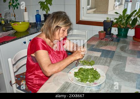 Eine ältere Frau saß an ihrem Küchentisch und Schnitt mit einem Messer vorsichtig grüne Bohnen. Die Szene spiegelt die Einfachheit und den Komfort des täglichen Lebens wider, wh Stockfoto