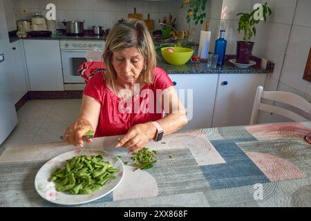 Eine ältere Frau saß an ihrem Küchentisch und Schnitt mit einem Messer vorsichtig grüne Bohnen. Die Szene spiegelt die Einfachheit und den Komfort des täglichen Lebens wider, wh Stockfoto