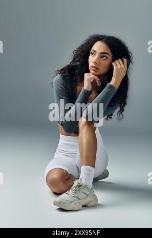 Eine athletische Afroamerikanerin sitzt auf einem grauen Hintergrund, schaut zur Seite und ruht sich nach einem Workout aus. Stockfoto