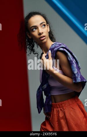 Eine junge Frau in einem Sport-BH und orangefarbenen Shorts lehnt sich in einem Studio an eine rote Wand mit farbenfrohen Glaspaneelen im Hintergrund. Stockfoto