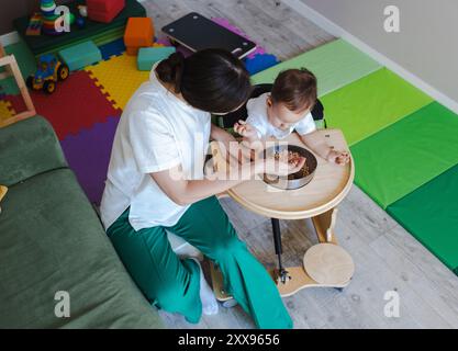 Ein kleiner Junge mit Zerebralparese betreibt mit einem Therapeuten eine körperliche Rehabilitation, umgeben von bunten Spielmatten und therapeutischen Werkzeugen. Stockfoto