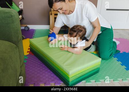 Ein kleiner Junge mit Hypotonie betreibt eine Physiotherapie, unterstützt von seiner Mutter in einem lebendigen Rehabilitationsraum Stockfoto