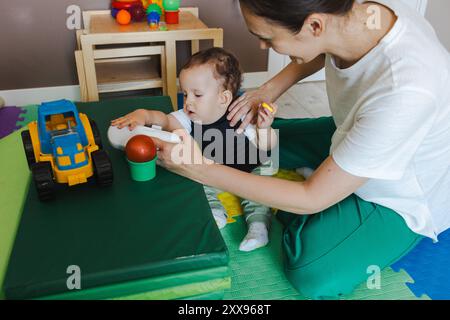 Junge mit Muskelschwäche nimmt Physiotherapie ein, während sie mit Spielzeug spielt, geführt von Physiotherapeuten in unterstützender Umgebung Stockfoto