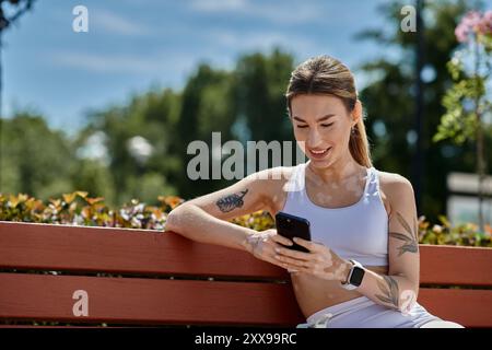 Eine junge Frau mit Vitiligo, die Aktivkleidung trägt, sitzt auf einer Bank in einem Park und lächelt, als sie ihr Handy überprüft. Stockfoto