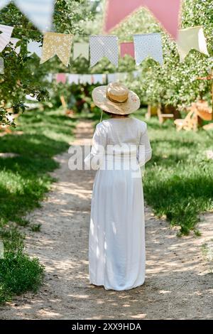 Eine Frau in weißem Kleid und Strohhut geht auf einem Weg durch einen Garten mit Lichterketten. Stockfoto