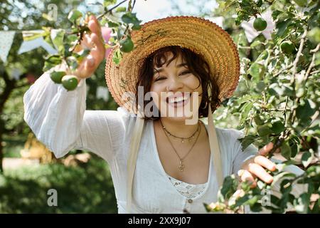 Eine junge Frau in weißer Bluse und Strohhut lächelt hell, während sie die Schönheit eines Sommergartens genießt. Stockfoto