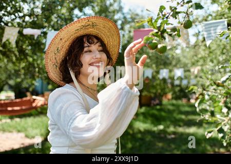 Eine junge Frau mit dunklem Haar, die einen Strohhut und eine weiße Bluse trägt, greift nach oben, um in einem üppigen Sommergarten Früchte von einem Baum zu pflücken. Stockfoto