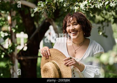 Eine junge Frau mit Strohhut und weißer Bluse lächelt hell, während sie in einem Sommergarten steht. Stockfoto