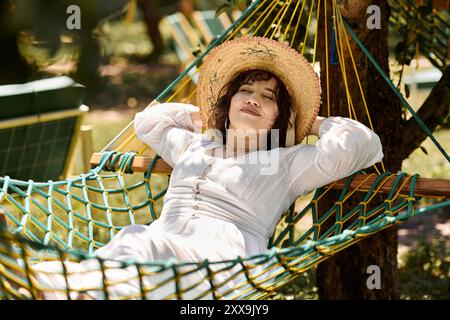 Eine junge Frau in weißem Kleid und Strohhut entspannt sich an einem sonnigen Sommertag in einer Hängematte. Stockfoto
