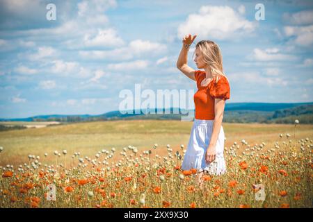 Blonde junge Frau, die in einem rot-weißen Kleid auf einem Feld mit rotem Mohn steht Stockfoto