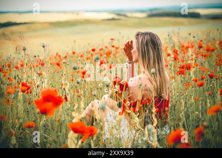 Blonde junge Frau in rot-weißem Kleid sitzt zwischen Blumen in einem Feld mit roten Mohnblumen Stockfoto
