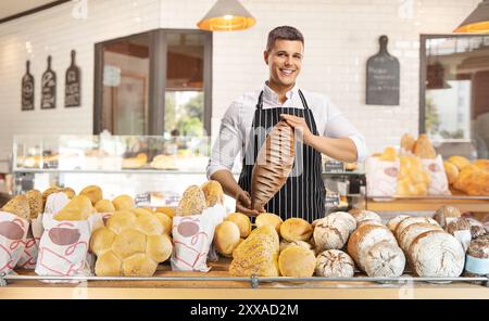 Junger, lächelnder Bäcker, der eine Schürze trägt und in einer Bäckerei einen Brotlaib hält Stockfoto