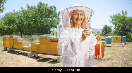 Imkerin in Schutzuniform, die Honig aus einem Glas probiert und lächelt Stockfoto