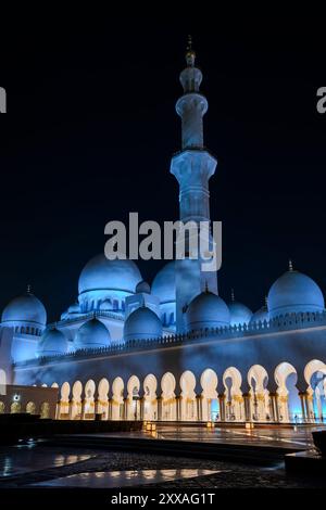 Seitenansicht der Scheich-Zayed-Moschee bei Nacht mit einigen der 82 weißen Marmorkuppeln, die von blauen sanften Lichtern beleuchtet werden, und eines der Minarette Stockfoto