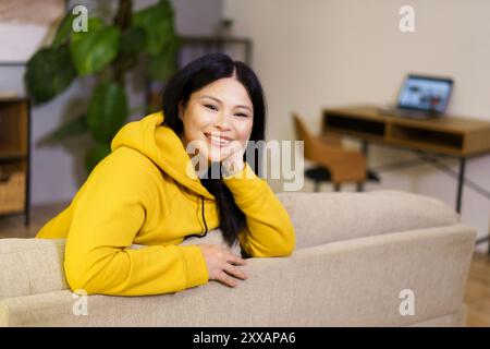 Eine Frau sitzt auf einer Couch und lächelt. Sie trägt einen gelben Hoodie. Das Zimmer verfügt über einen Laptop auf einem Schreibtisch und eine Topfpflanze Stockfoto