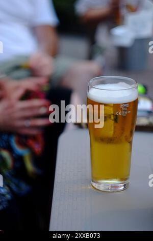 Ein halbes Glas Bier mit Kondenswasser steht auf einem Tisch vor einem Restaurant mit Leuten, die an einem Tisch im Hintergrund sitzen Stockfoto
