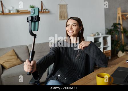 Eine junge Frau in einem grauen Pullover sitzt an einem Schreibtisch in ihrem Haus und filmt mit einem Selfie-Stick Inhalte auf ihrem Handy. Stockfoto